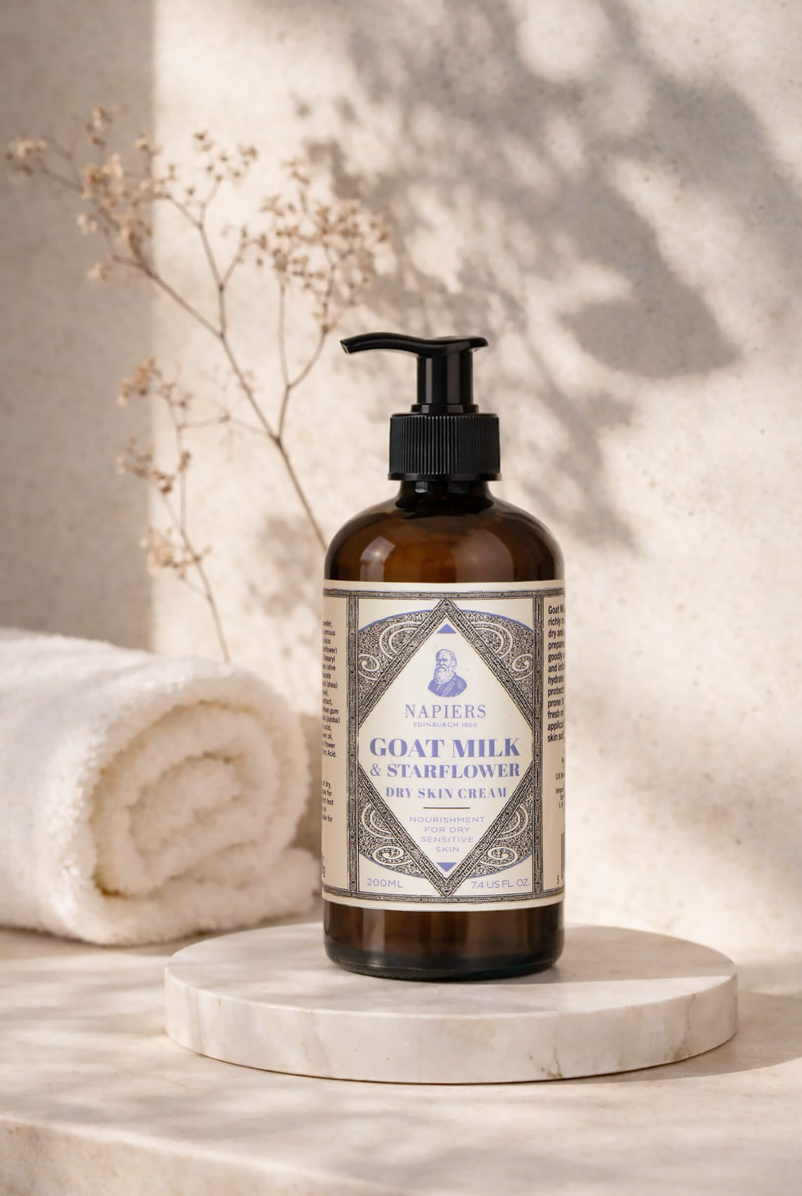 Napiers Goat Milk & Starflower Dry Skin Cream on marble pedestal with rolled towel and dried flowers